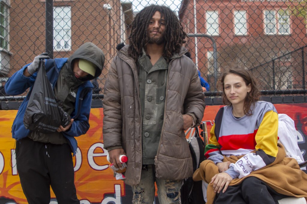 Three people, two young men and a woman in a wheelchair, pose in front of a fence.