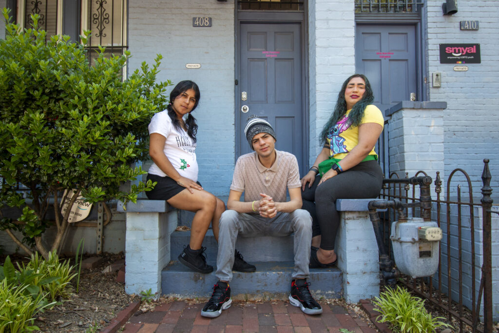 Three Latino youth pose in front of a blue building, looking at the camera.