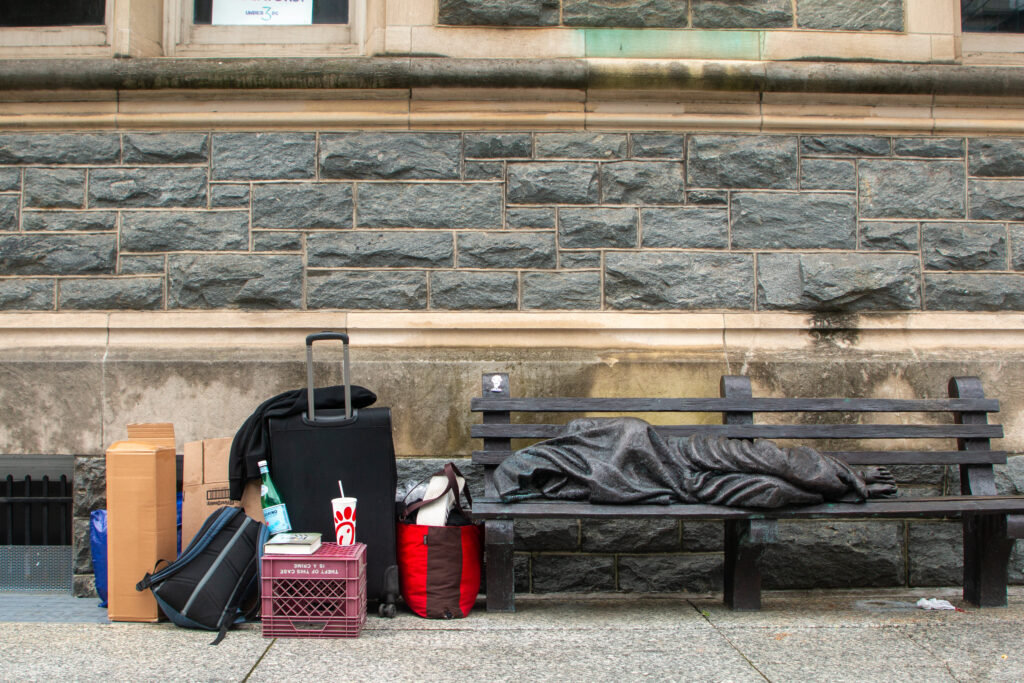 Bags and suitcases to the left of a statue depicting a homeless man sleeping on a bench