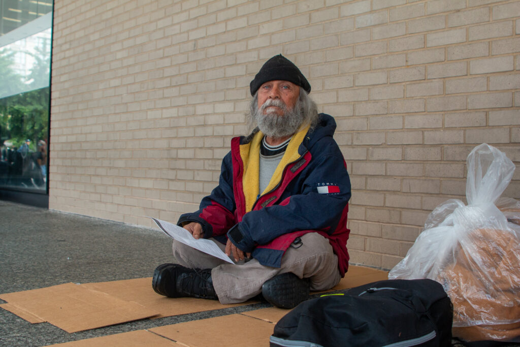 A man in a blue and yellow jacket sits on a piece of cardboard in front of a stone wall.