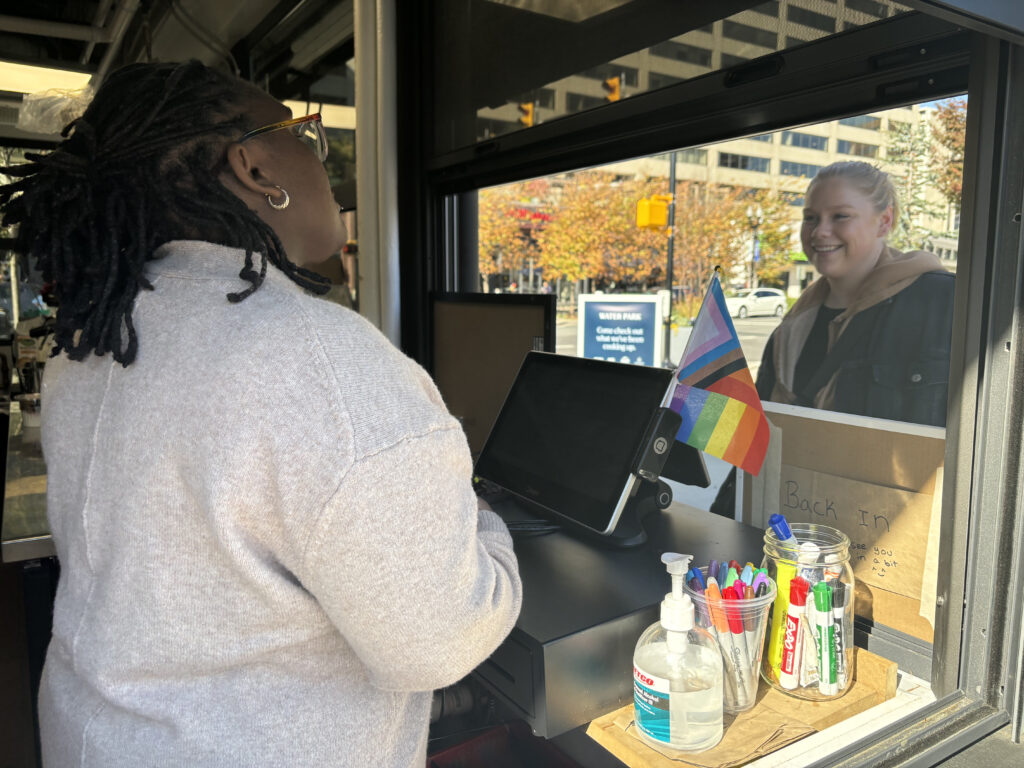 A Black woman inside a coffee stand talks to a customer.