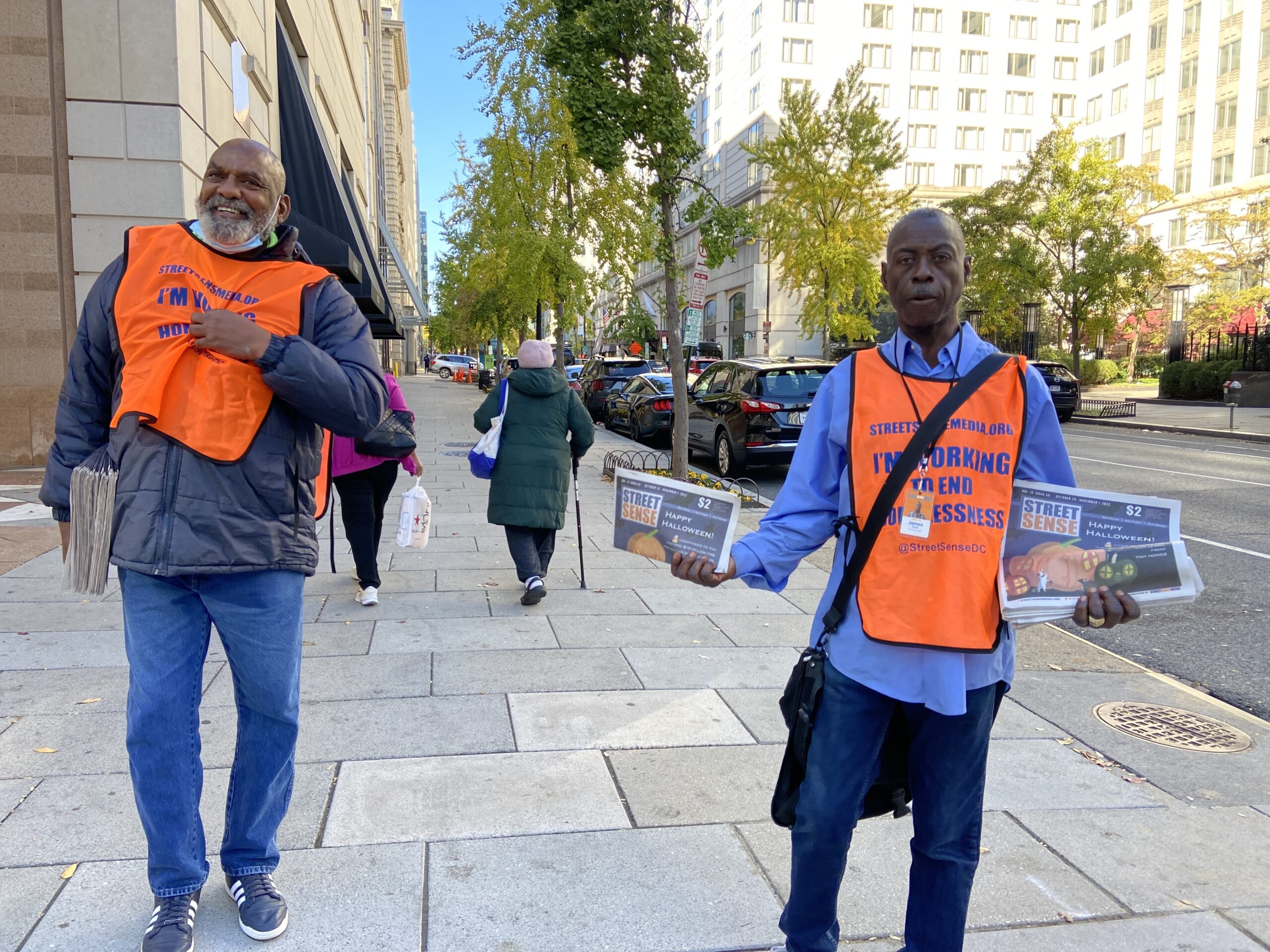 Two smiling men wearing Street Sense Media vests sell papers.