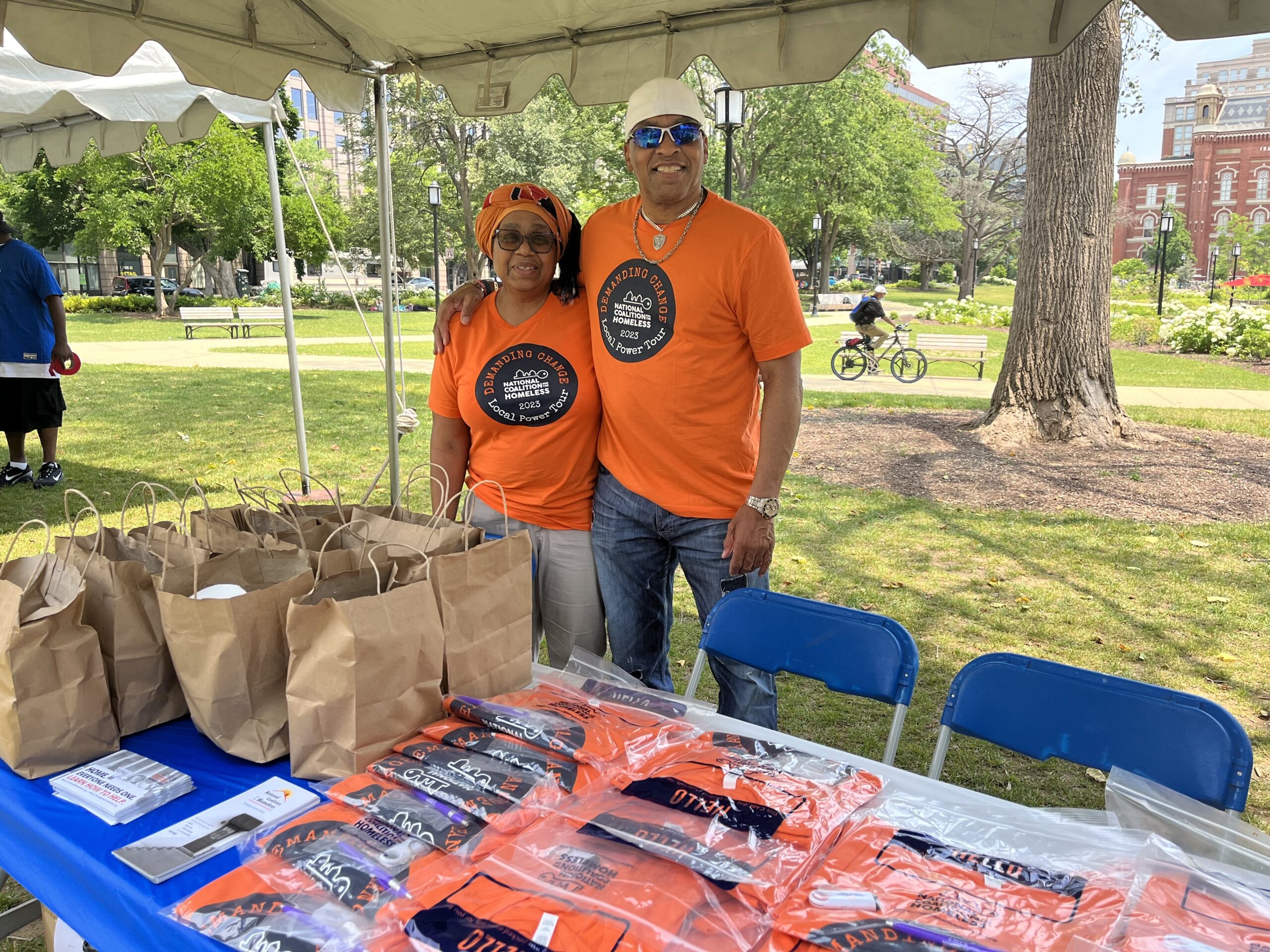 Two people stand side-by-side in orange shirts in front of a table with fliers and gift bags.