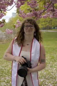 A person stands in graduation regalia in front of a tree.