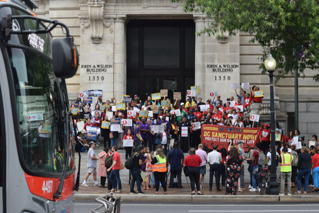 A group of people wearing colorful shirts and holding signs about the budget stand in front of D.C.'s city building.