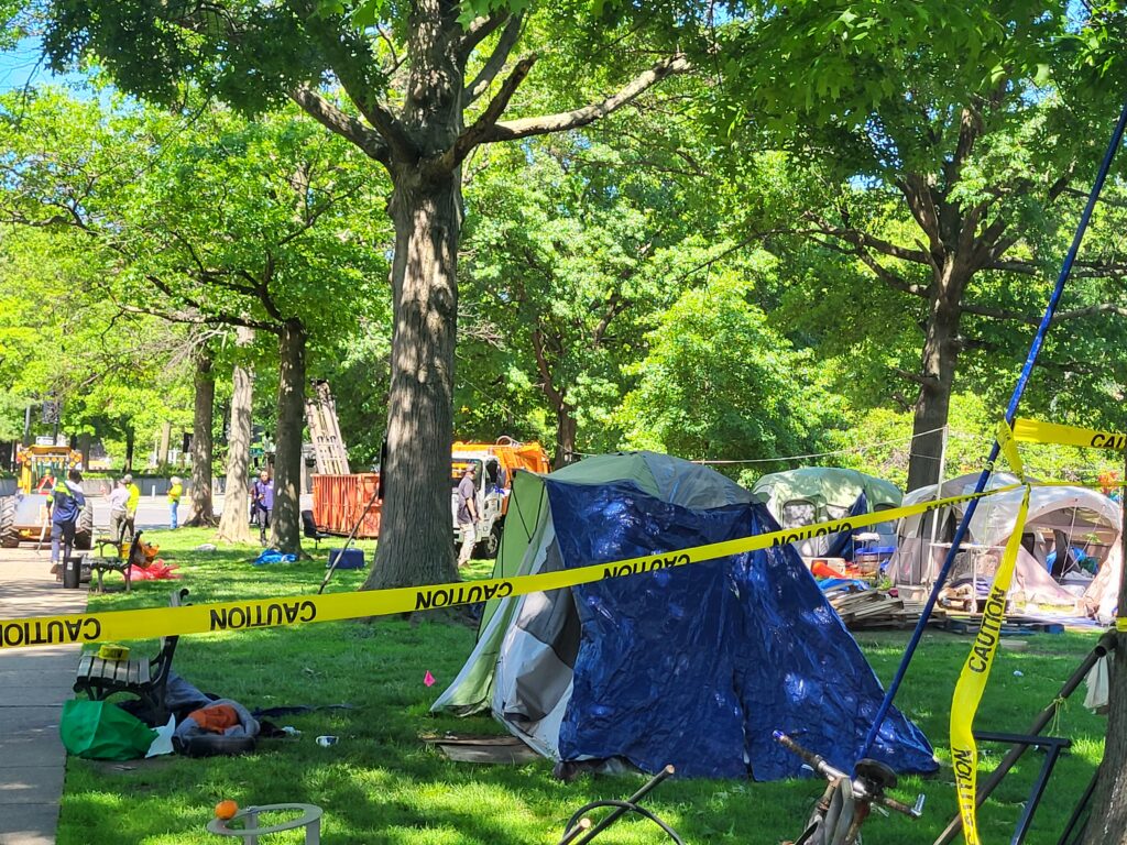 A blue tent against green trees and grass, with caution tape in front of it.