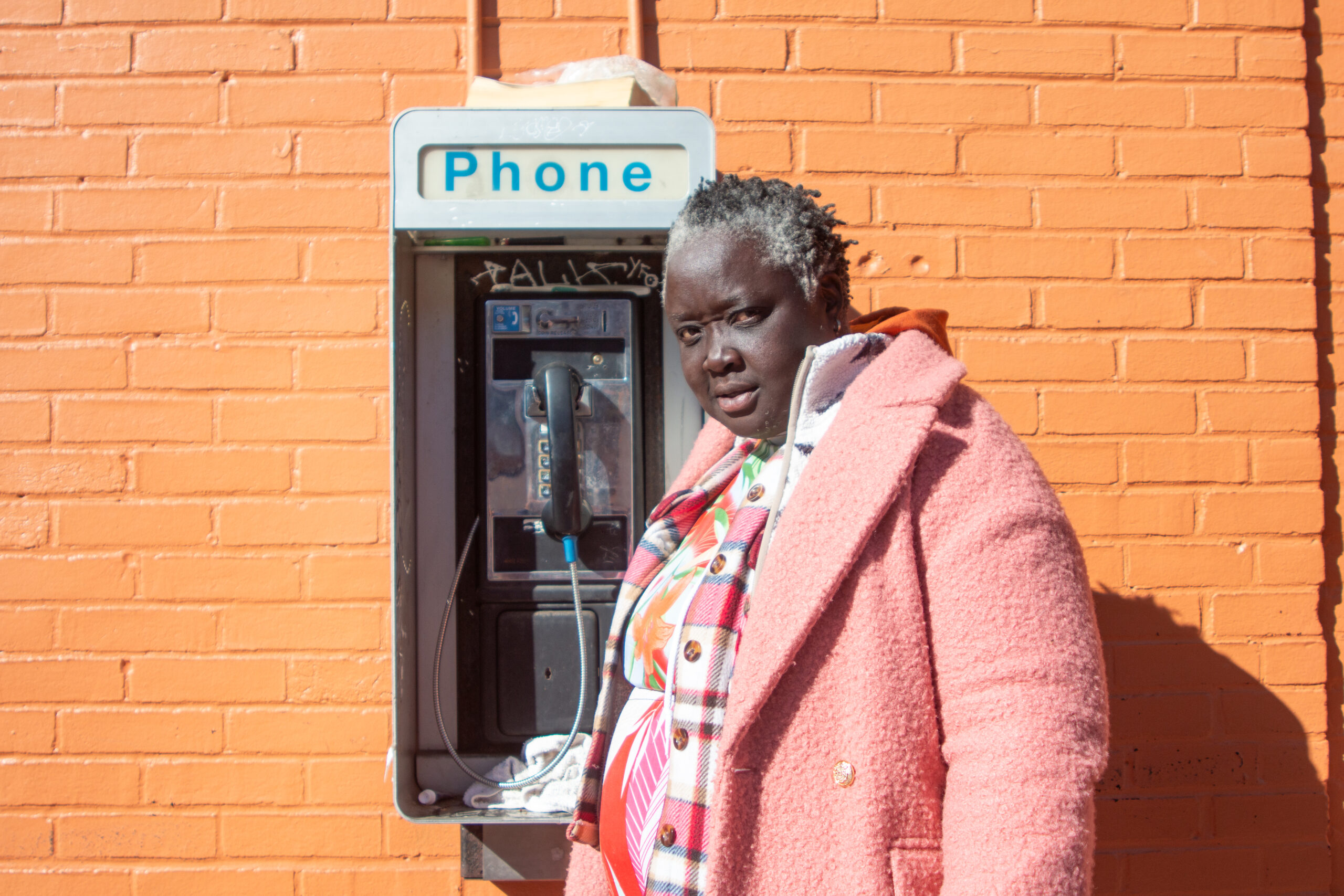 A Black woman in a pink coat stands against an orange wall near her encampment