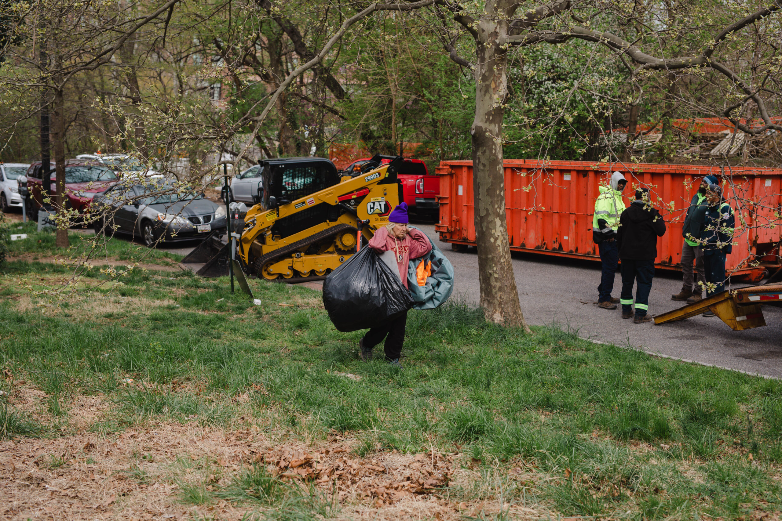A white woman in a pink shirt carries two black trash bags across grass.