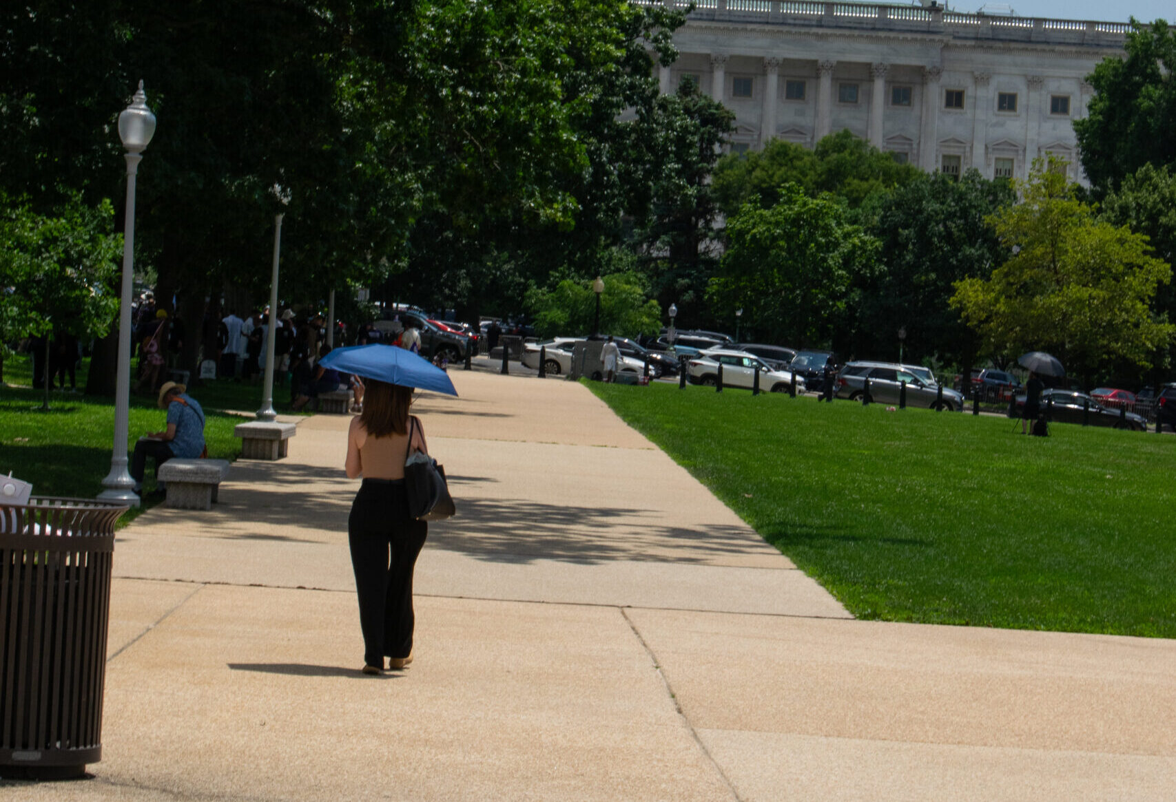 A woman under a blue umbrella walks to the Capitol on a sunny day.