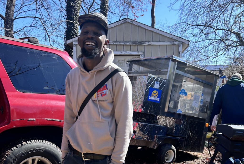 A Black man stands smiling in front of a food cart.