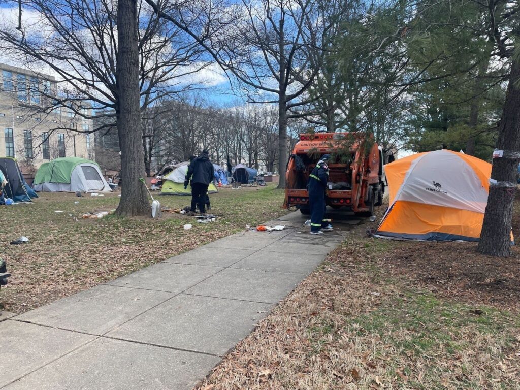 Officials in dark clothing throw away trash near tents in a park.
