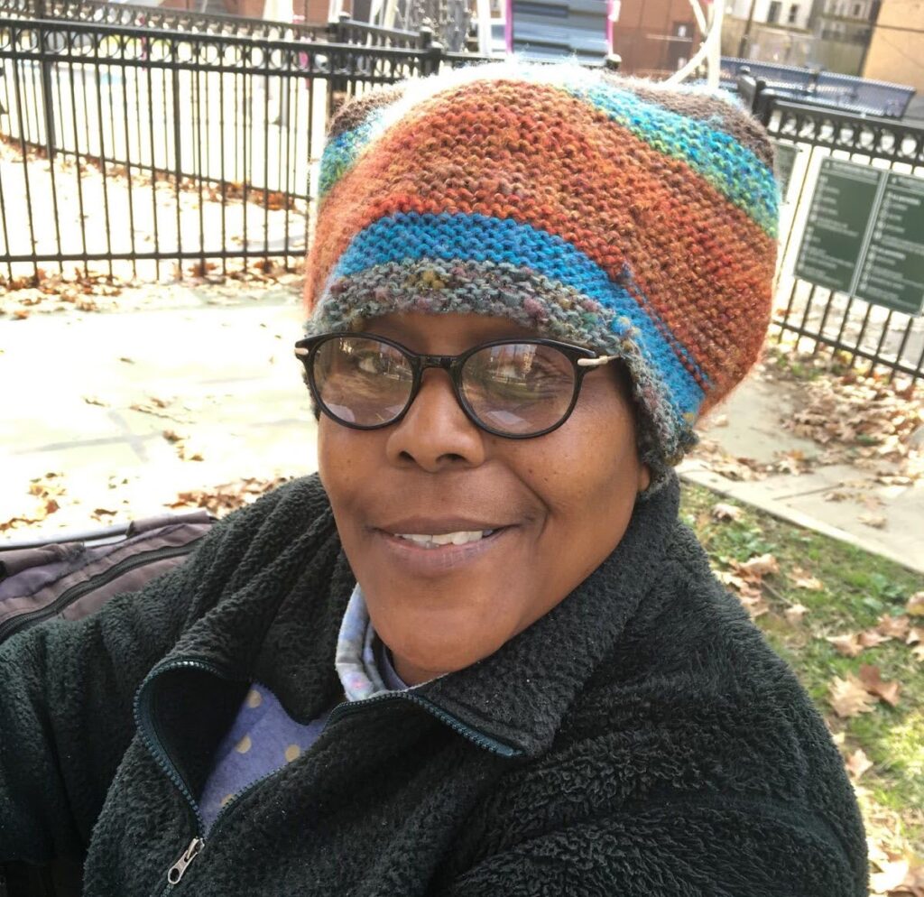 A Black woman in a knitted hat smiles up at the camera.