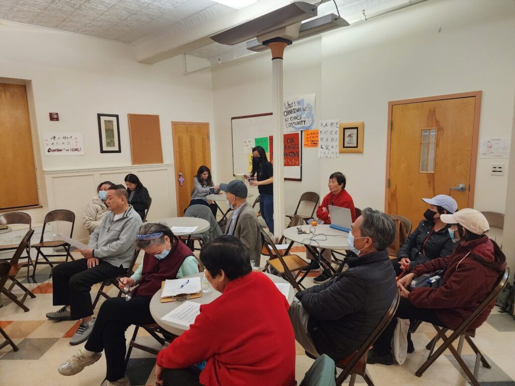 A group of Chinatown residents sit in folding chairs in a community room.