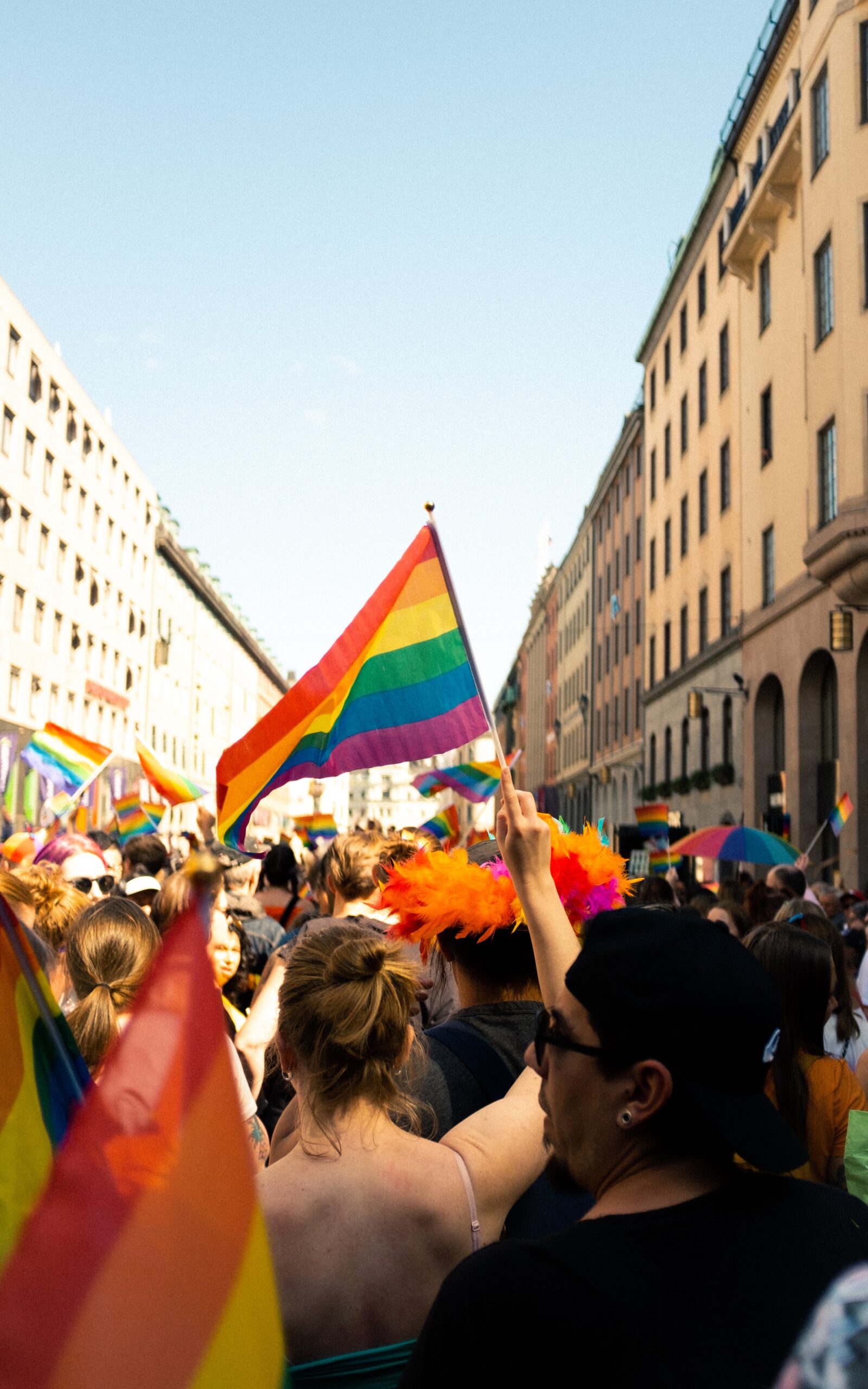 A blond woman holds a pride flag with a parade behind her.